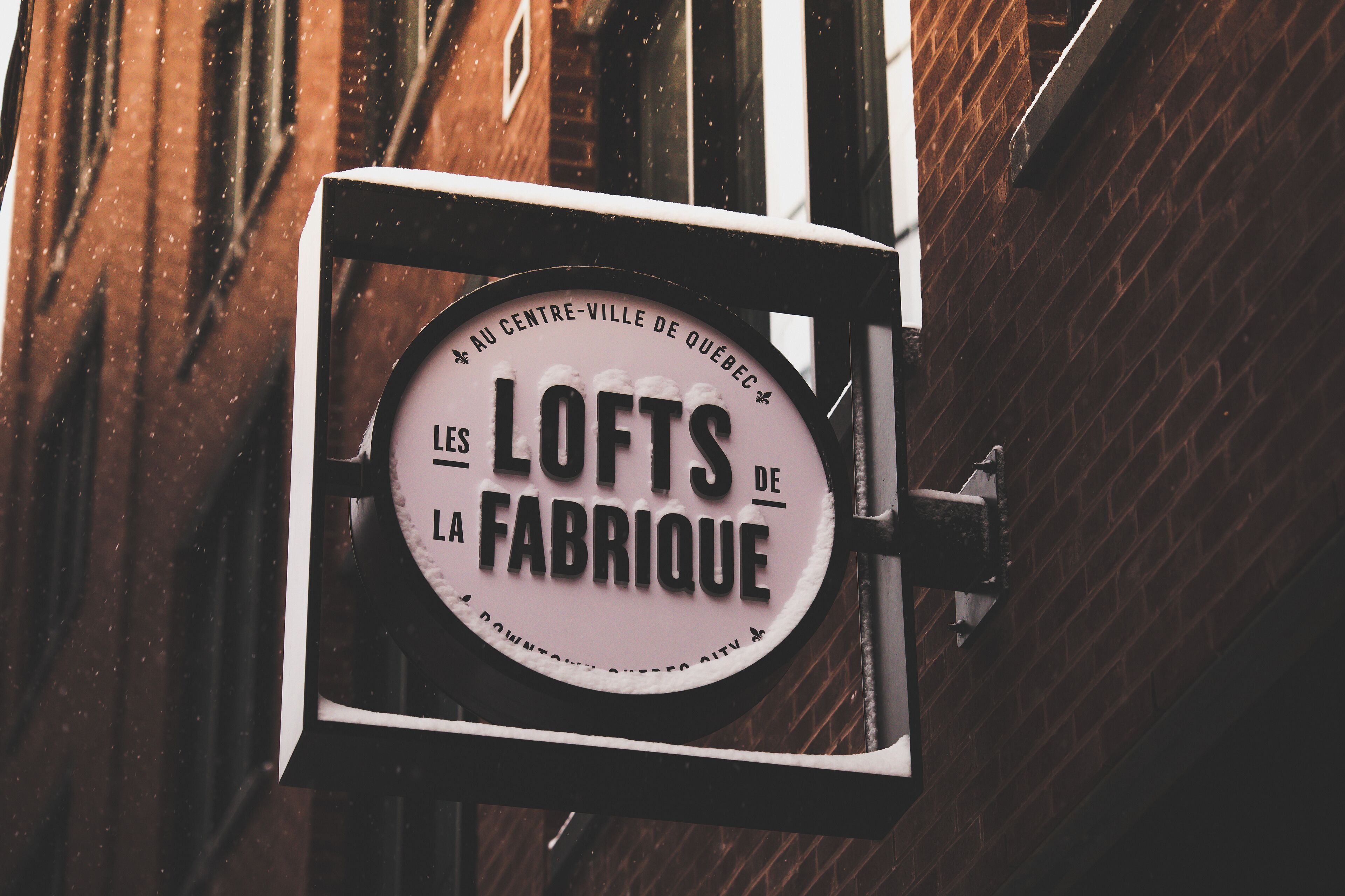 A sign with the text "LES LOFTS DE LA FABRIQUE" mounted on a metal frame against a brick wall.
