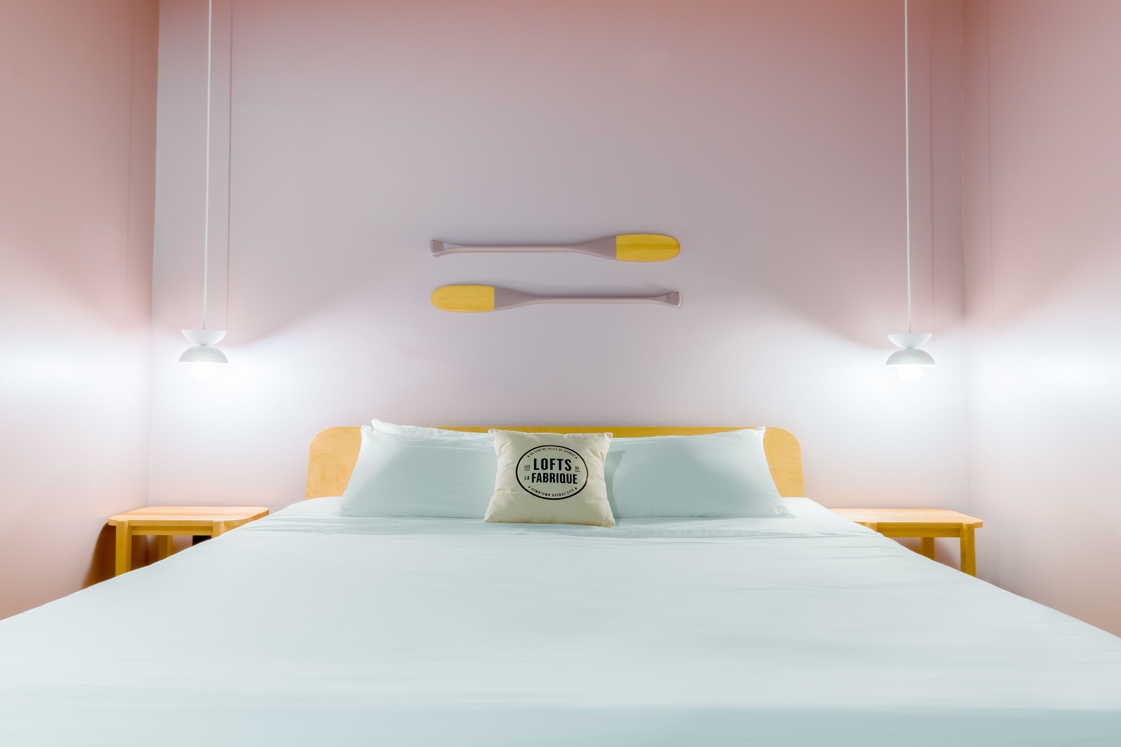 Ein ordentlich bezogenes Bett mit weißer Bettwäsche und einem Kissen mit einem Logo, zwei Nachttische und eine an der Wand befestigte Leuchte.