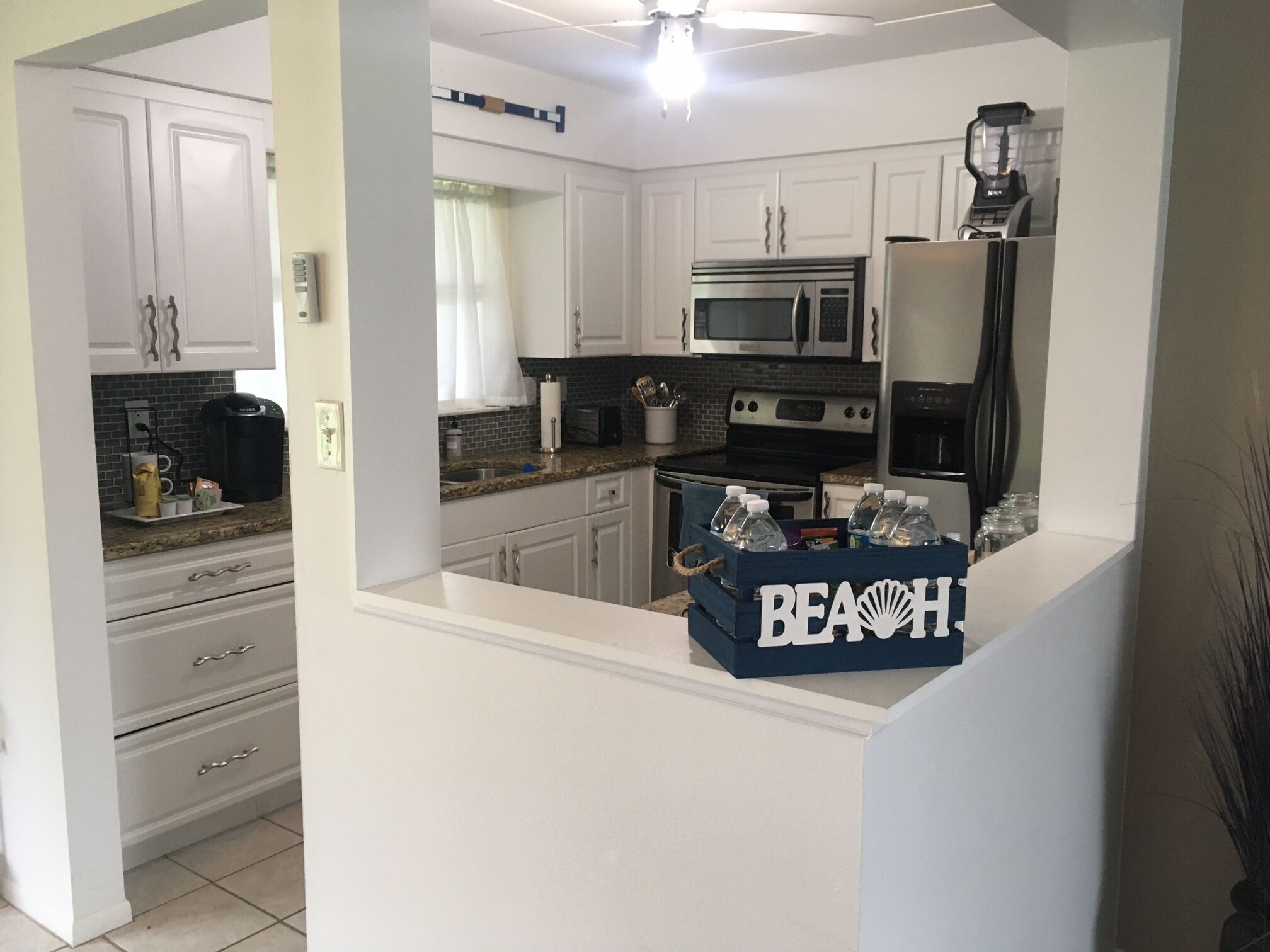 A modern kitchen with white cabinets, a granite countertop, a microwave, an oven, and a refrigerator. There is a blue box labeled "BEACH" on the counter.
