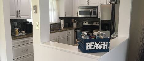 A modern kitchen with white cabinets, a granite countertop, a microwave, an oven, and a refrigerator. There is a blue box labeled "BEACH" on the counter.