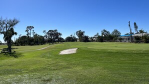 Un campo de golf con cielo despejado, árboles y un edificio al fondo.