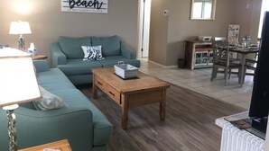 Una sala de estar con un sofá verde, una mesa de centro de madera y un comedor con mesa y sillas. Un letrero en la pared dice "The beach is my happy place".