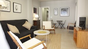 A living room with a black sofa, a round coffee table, and a TV stand. There are framed pictures on the walls and a lamp on a side table.