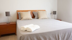 A neatly made bed with a grey bedspread and two pillows, a wooden nightstand with a lamp, and a wall socket.