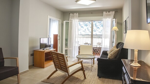 A living room with a TV, a sofa, a chair, a small table, and a lamp. There is a balcony with a view of the sky.