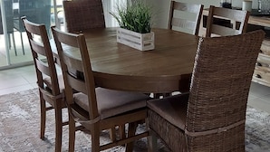 Een eetruimte met een ronde houten tafel, rieten stoelen, een kroonluchter en uitzicht door grote ramen.