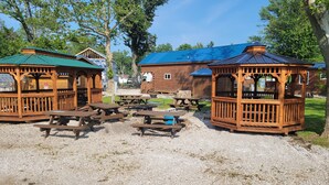 Un chalet rustique en bois, doté d’un toit vert, entouré de tables et de bancs de pique-nique, situé dans un espace recouvert de gravier, avec des arbres en arrière-plan.