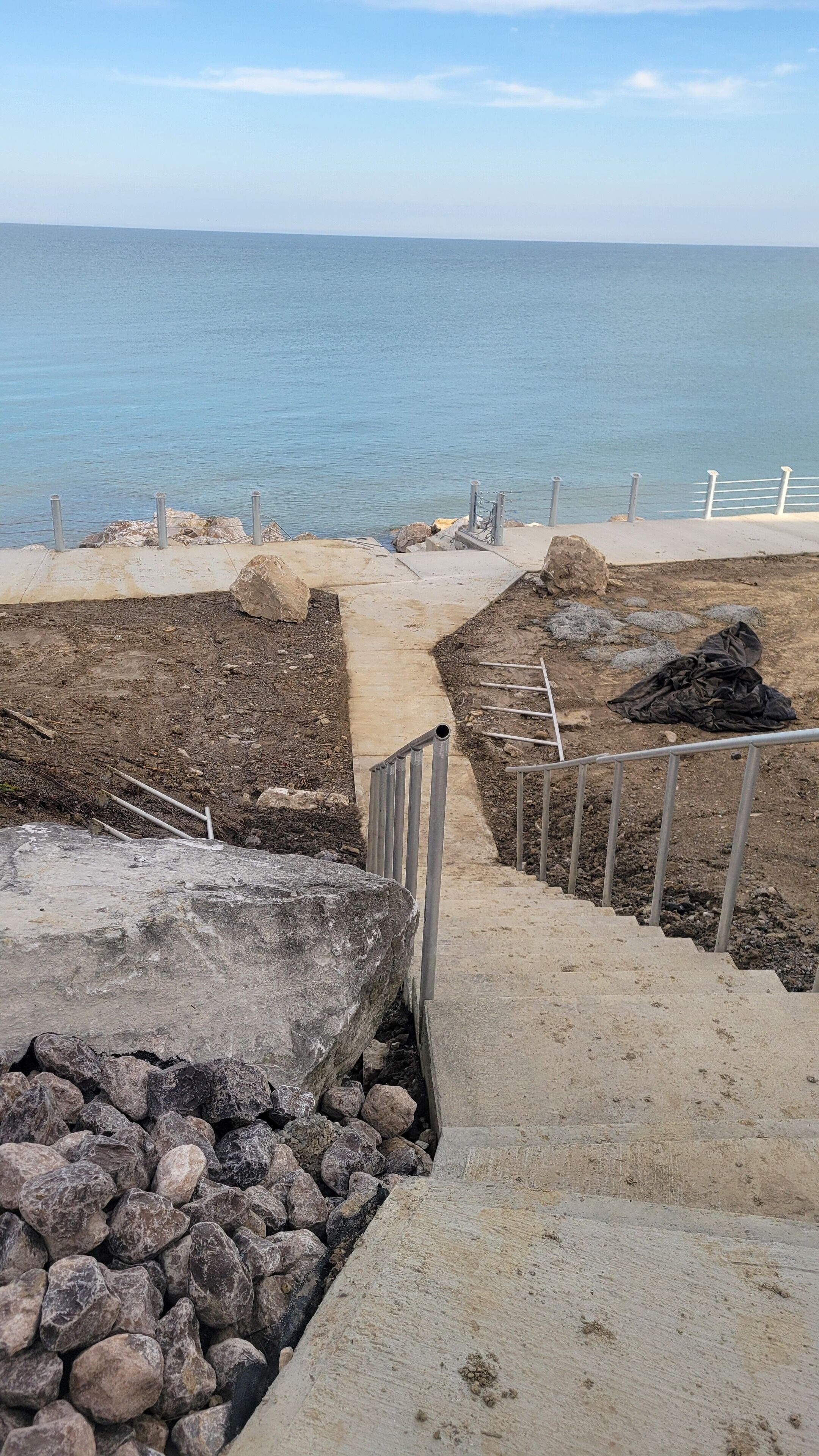 Une promenade en bord de mer, avec des marches en béton menant à une zone sablonneuse, des garde-corps en métal et une vue sur la mer.