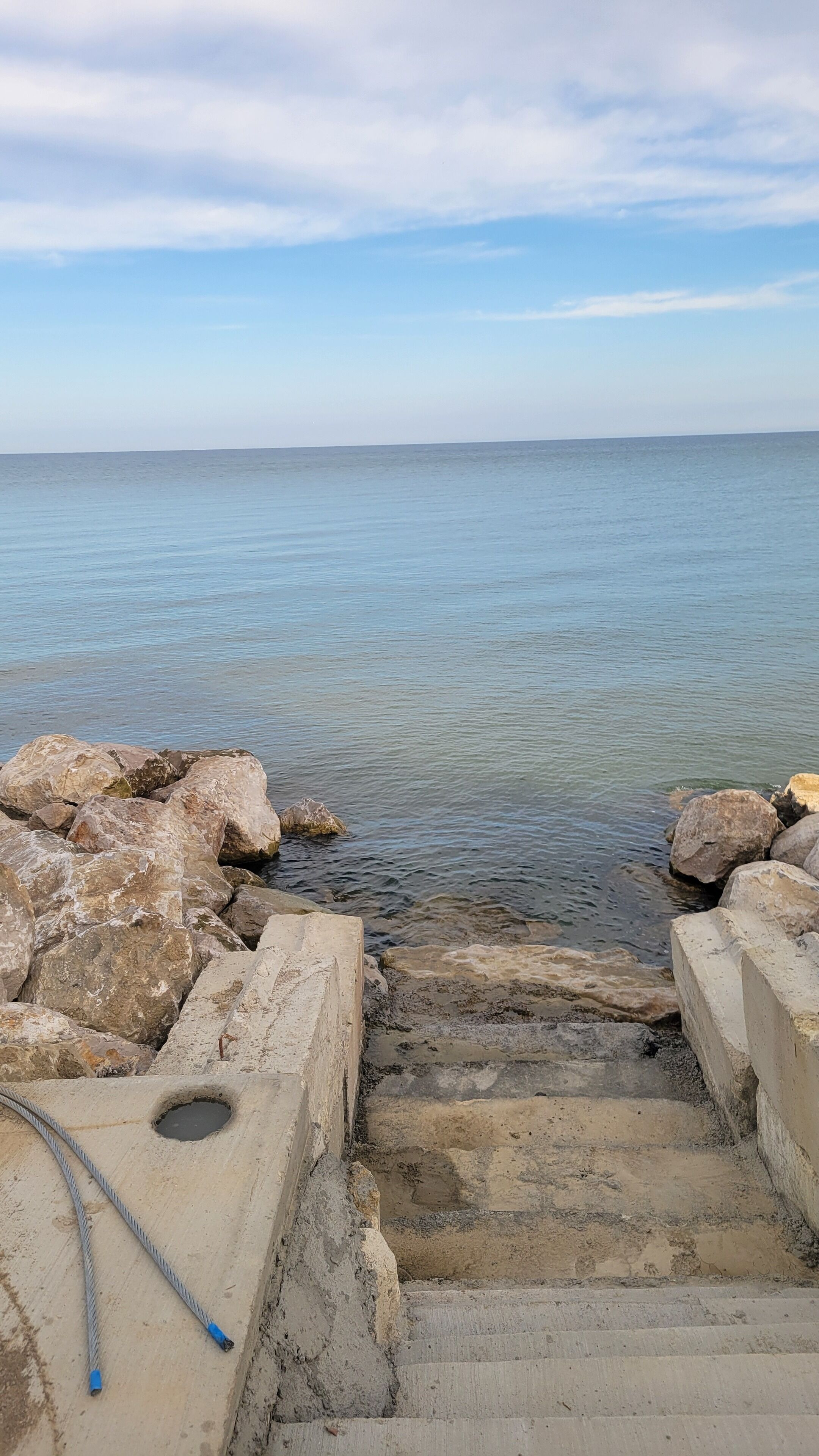 Una escalera de hormigón que desciende hacia un cuerpo de agua, rodeada de grandes rocas y un cielo despejado.