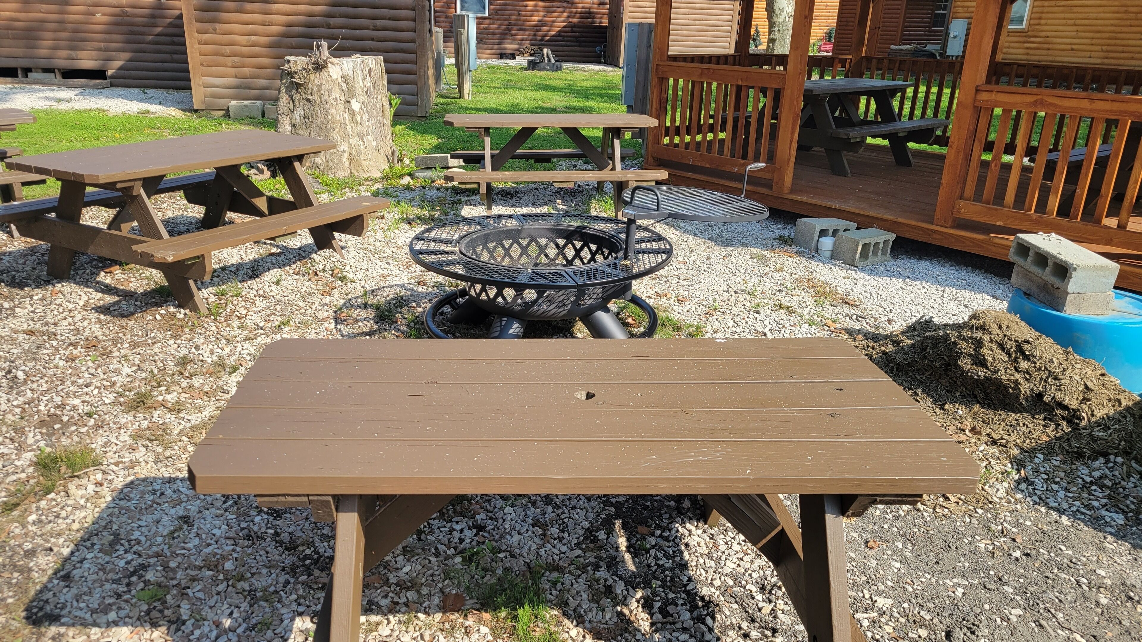 A wooden picnic table with a metal grill in the center, surrounded by gravel and grass.