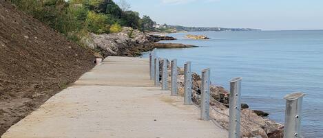 A concrete pathway along a rocky shoreline with metal posts and a clear view of the sea.