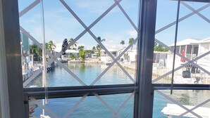 Vista desde una ventana con celosía metálica, que da a un puerto deportivo con barcos y un muelle. En la terraza hay sillas de mimbre y una pequeña mesa.