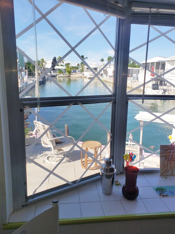 A view from a window with a metal grid, overlooking a marina with boats and a dock. There are wicker chairs and a small table on the deck.