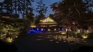 Una casa con techo triangular y varias ventanas, rodeada de árboles e iluminada por luces.