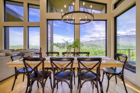 Uma área de refeições com mesa e cadeiras de madeira, grandes janelas com vista para as montanhas e um lustre.