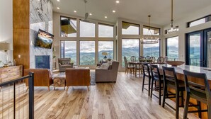 A spacious living area with a large window overlooking a mountainous landscape, a dining area with a long table and chairs, and a cozy seating area with a sofa and armchairs.