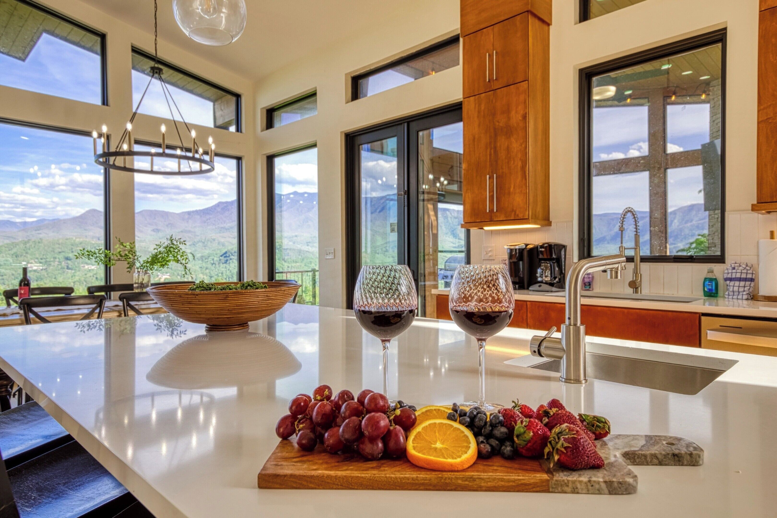 A kitchen with a large island, a bowl of fruit, two wine glasses, and a view of mountains.