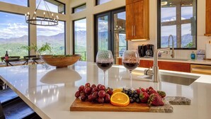 A kitchen with a large island, a bowl of fruit, two wine glasses, and a view of mountains.