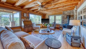 L’intérieur chaleureux d’un chalet avec un plafond en bois, une cheminée en pierre, un canapé, une table à manger et une grande fenêtre donnant sur la forêt.