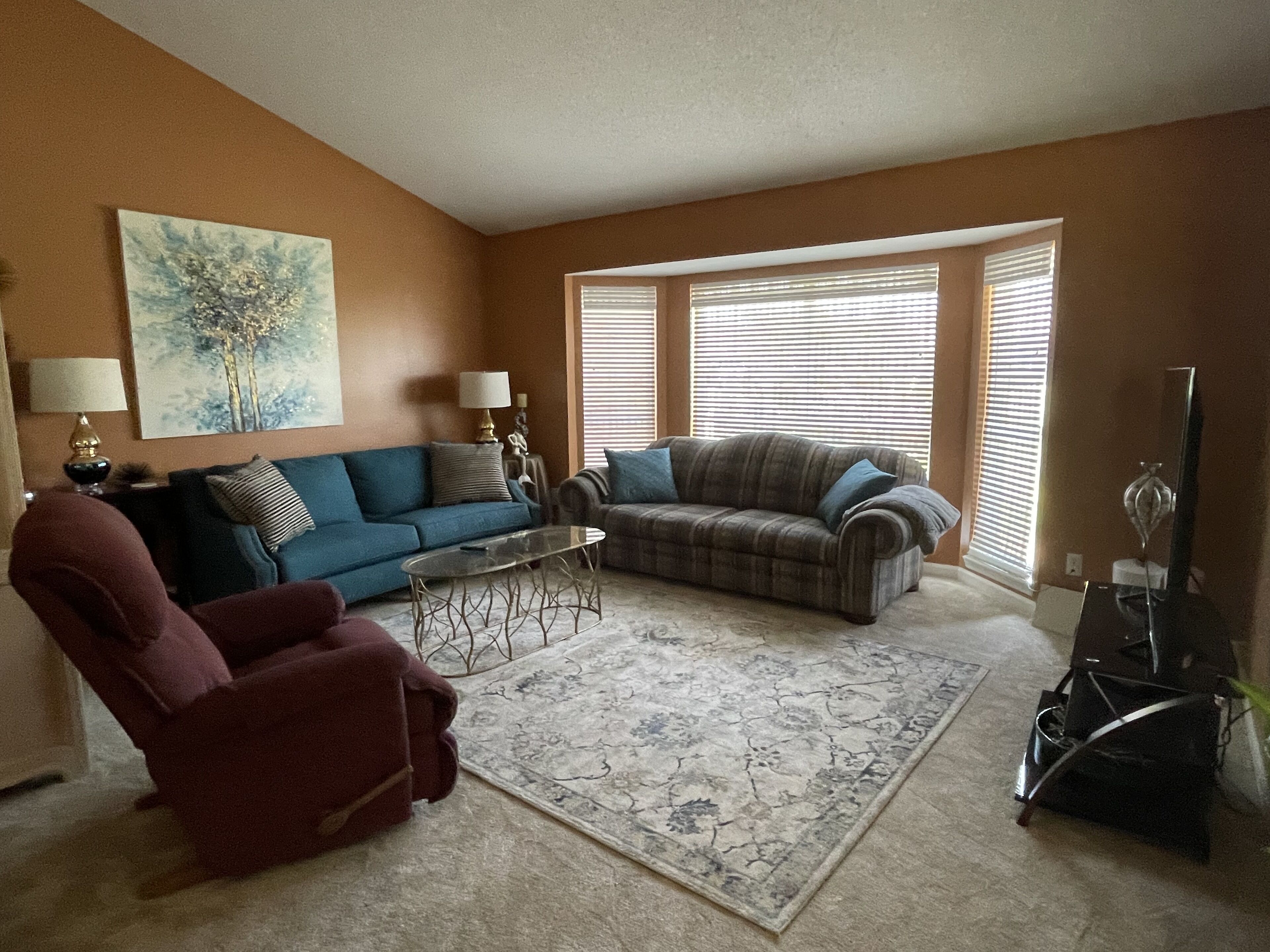 A living room with two sofas, a coffee table, a red armchair, and a large window with blinds.