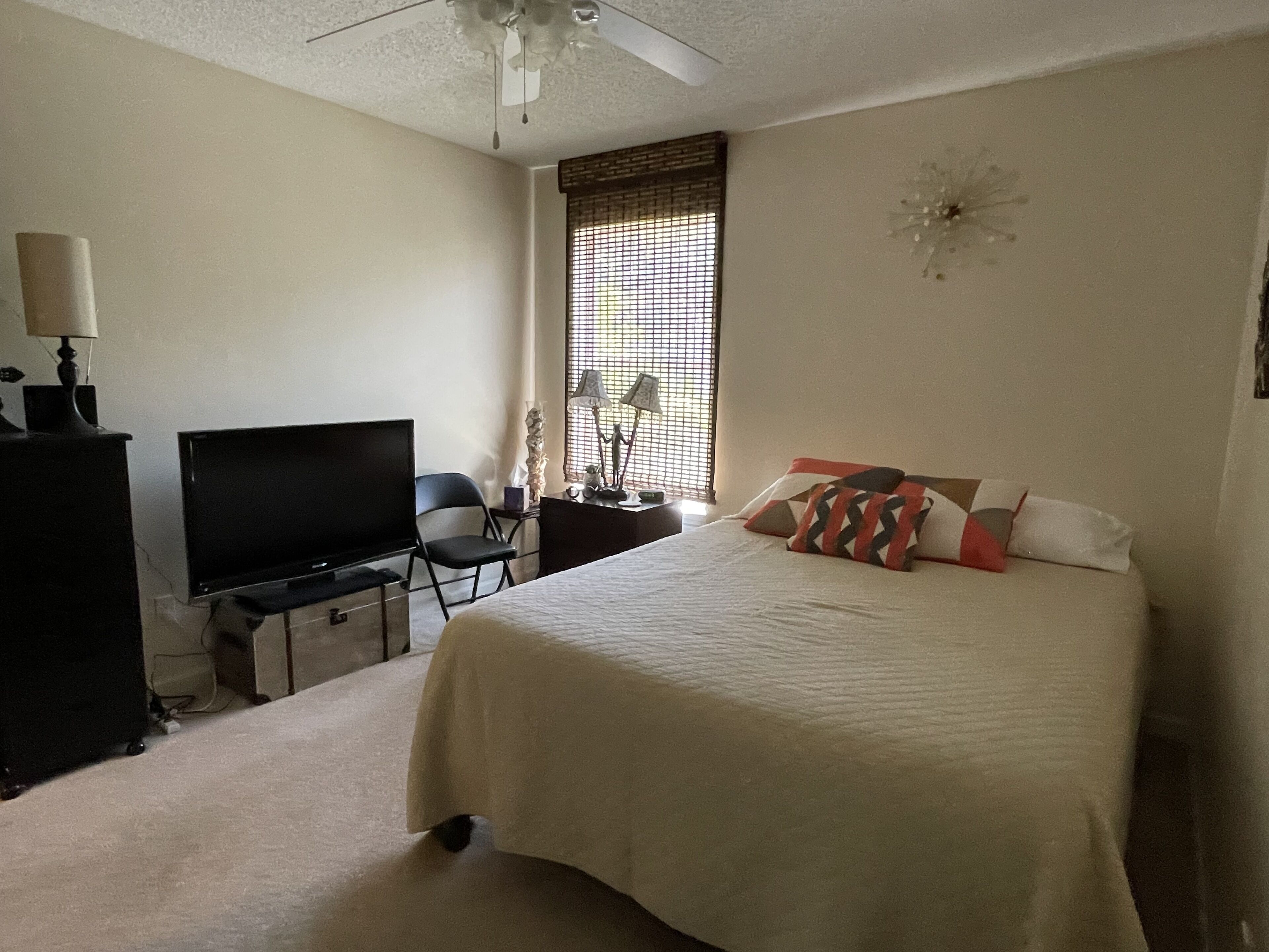 A bedroom with a bed, a desk, a chair, a television, and a window with blinds.