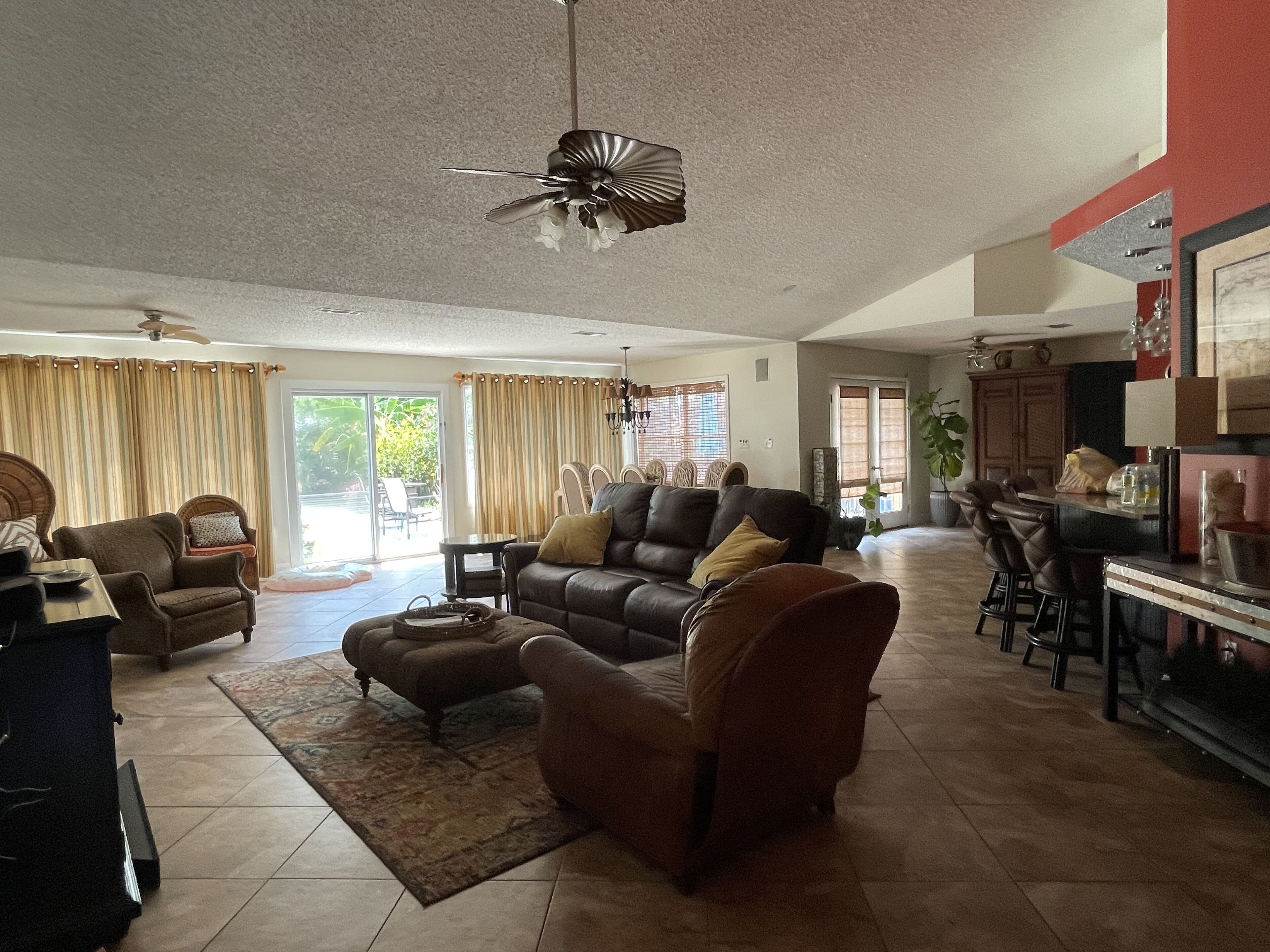 A spacious living room with a sofa, armchairs, and a coffee table. There is a dining area with a bar and chairs. The room has large windows with curtains and a ceiling fan.