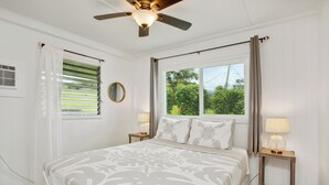 Ein Schlafzimmer mit Bett, Deckenventilator, Fenster mit Vorhängen, Nachttisch mit Lampe und einem wandmontierten Spiegel.