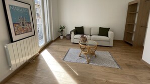 Una sala de estar con un sofá blanco, una mesita de mimbre, un cuadro enmarcado, un radiador y una estantería de madera.