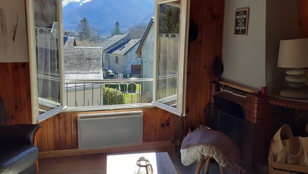 Une pièce chaleureuse avec une grande fenêtre donnant sur une montagne et des bâtiments. On y trouve une cheminée, une table avec une bougie et un fauteuil.