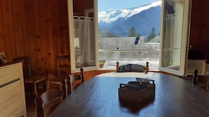 Une table à manger en bois, des chaises, une fenêtre avec des rideaux et une vue sur des montagnes enneigées.
