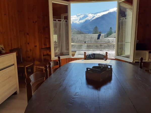 A wooden dining table with chairs, a window with curtains, and a view of snow-capped mountains.