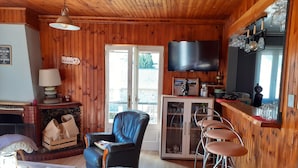 L’intérieur chaleureux d’un chalet avec un plafond en bois, une cheminée, un téléviseur fixé au mur et un coin bar avec des tabourets.