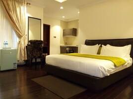 Quarto de hotel com uma cama grande, uma escrivaninha, uma cadeira, um espelho e uma cozinha compacta.