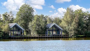 Dues cabanes modernes amb grans finestres, ubicades sobre un moll de fusta a un llac tranquil, envoltades per arbres densos.