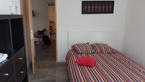 A bedroom with a bed, a red pillow, a striped blanket, a black wardrobe, and a framed Coca-Cola bottle artwork on the wall.