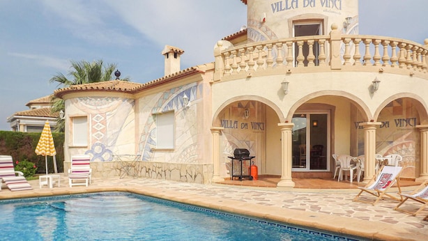 Zona de piscina amb gandules i ombrelles, edifici amb el rètol 'Villa da Vinci' i solera de piscina amb rajoles.