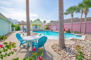 A backyard with a swimming pool, a thatched-roof structure, and a pink building.