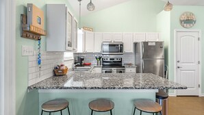 Una cocina con armarios blancos, encimera de granito, refrigerador de acero inoxidable y microondas. Hay tres taburetes en la isla.