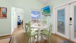 Un comedor con una mesa redonda y cuatro sillas, un televisor montado en la pared, y una puerta con mosquitero.