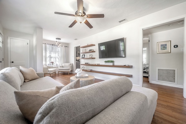 Un salon moderne doté d’un ventilateur de plafond, d’un téléviseur à écran plat et d’un espace détente avec un canapé et des fauteuils.