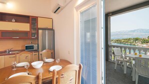 Spiseområde med et bord dækket til fire personer, et køkken med køleskab og mikroovn, samt en balkon med udsigt over havet og bjergene.