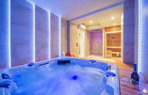 A modern spa room with a large hot tub, a sauna, and a wooden massage table.