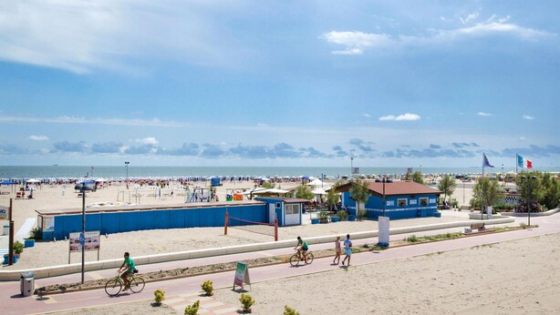 Une zone située au bord de la plage, avec un bâtiment bleu portant l’inscription « CAFÉ, RESTAURATION RAPIDE, BOISSONS », une plage de sable et une route où l’on peut voir des cyclistes.