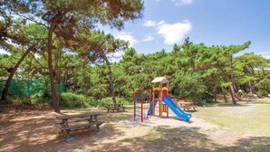Une aire de jeux avec un toboggan et des structures d’escalade, entourée d’arbres et de tables de pique-nique.