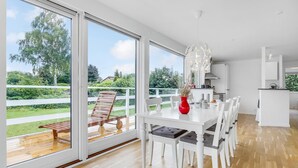 A modern kitchen with white cabinets and a large island, a dining area with a white table and chairs, and a sliding glass door leading to a deck with a wooden bench. - Beautiful home in Strøby with house sea view (Strøby)