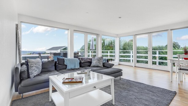 A modern living room with a grey sofa, a glass coffee table, and large windows offering a view of the sea and greenery. - Beautiful home in Strøby with house sea view (Strøby)