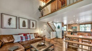 A spacious living area with a high ceiling, a wooden staircase, a dining area with a long table, and a kitchen with modern appliances.