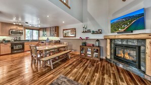 A spacious kitchen with wooden flooring, a dining area with a large wooden table and benches, a fireplace, and a mounted TV displaying a scenic view.