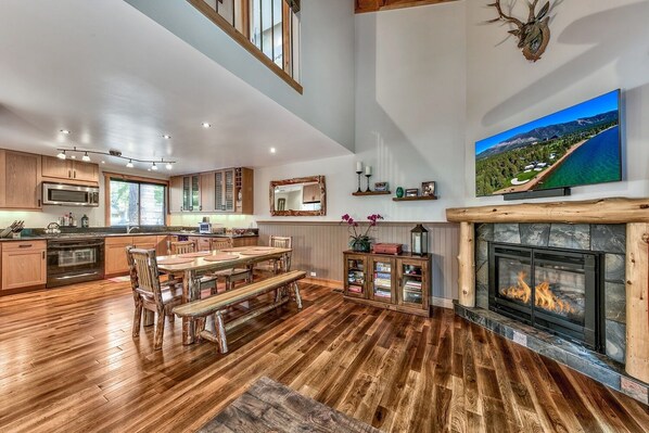 Una cocina amplia con piso de madera, un comedor con una gran mesa de madera y bancos, una chimenea y un televisor montado que muestra una vista panorámica.