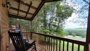 A wooden deck with a rocking chair and a table, overlooking a forested area with a view of distant hills. - "The DogHouse" a Pet Friendly Cabin on a 70 ac horse farm near Fall Creek Falls! (Van Buren County)